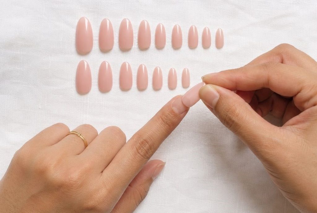 A flat lay view of neutral pink press-on nails organized by size, with hands checking for the correct fit